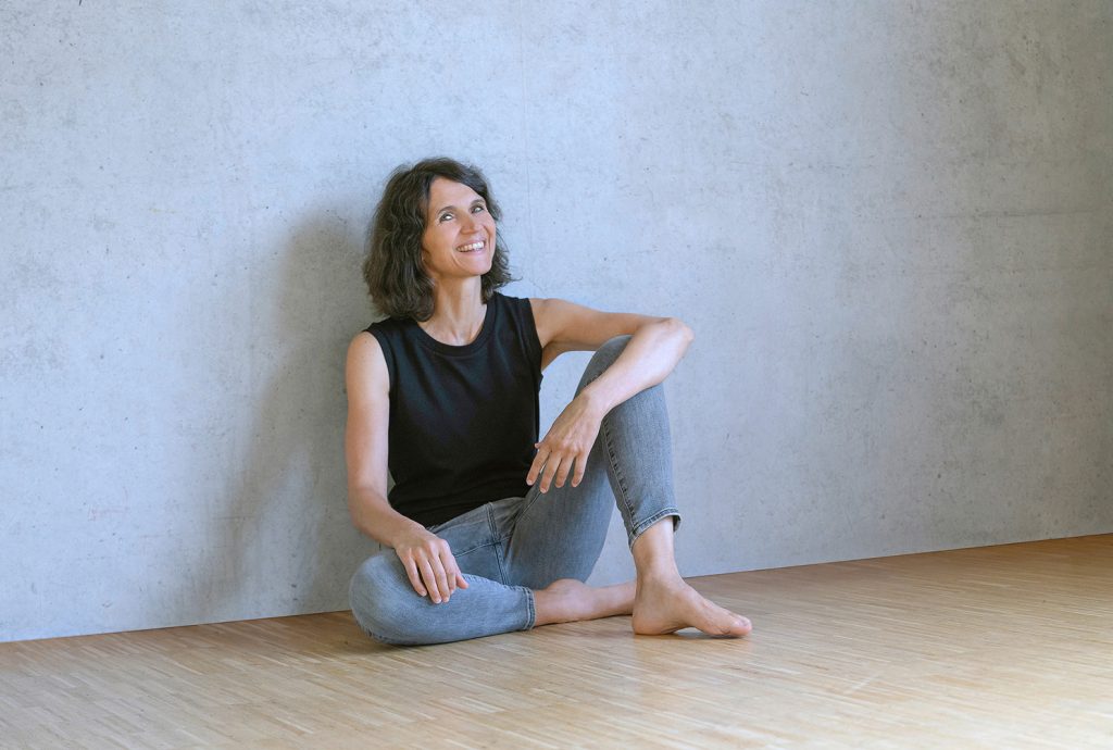 Sandra Schnetzer, Yoga-Lehrerin bei Yoga Blue in Stuttgart, sitzt auf einem Holzboden vor einer Betonwand und strahlt Ruhe und Gelassenheit aus.